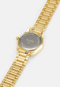 Gouden roestvrijstalen horloge met een schakelband en een ronde kast. De achterkant heeft gegraveerde tekst op een glad oppervlak, dat de waterbestendigheid aangeeft.