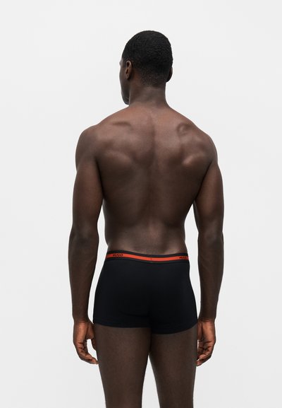 Homme à la peau foncée et aux cheveux courts vu de dos, portant un boxer noir avec une ceinture rouge et le mot "HUGO" dessus.