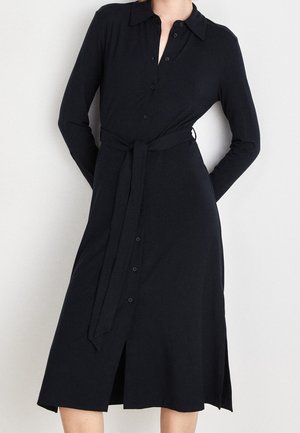 Femme portant une robe noire à manches longues, boutonnée, avec un col et une ceinture nouée à la taille, debout devant un fond uni.