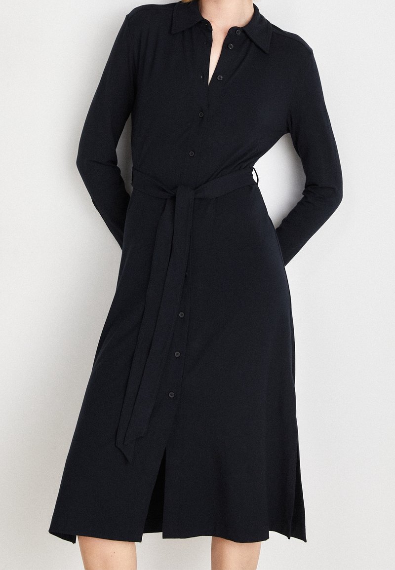 Femme portant une robe noire à manches longues, boutonnée, avec un col et une ceinture nouée à la taille, debout devant un fond uni.