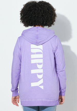 Persona con capelli ricci che indossa una felpa con cappuccio viola chiaro con la scritta bianca verticale "HAPPY" sulla schiena, in piedi davanti a uno sfondo bianco.