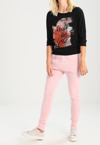 Schwarzes Langarm-Oberteil mit Textdesign, kombiniert mit hellrosa Jogginghosen. Weiße Turnschuhe runden das Outfit ab. Flache, glatte Texturen sind sichtbar.