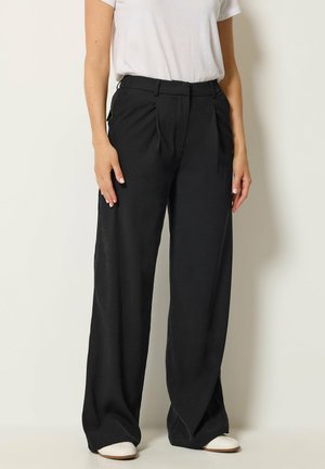 Person trägt ein weißes Kurzarm-T-Shirt, hochgeschnittene schwarze weite Hosen und weiße Schuhe, stehend vor einer schlichten Wand.