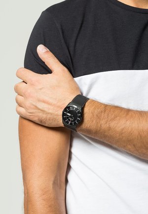 Schwarz gehaltene Uhr mit einem strukturierten Mesh-Armband, rundem Zifferblatt und silbernen Stundenmarkierungen, getragen an einem nackten Arm vor einem schwarz-weißen Hemd.