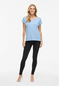 T-shirt azzurro chiaro a maniche corte, vestibilità comoda, abbinato a jeans skinny neri e sandali con tacco bianchi, su uno sfondo bianco uniforme.