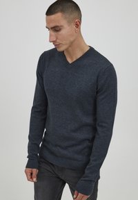 Maglione blu a scollo a v realizzato in tessuto lavorato a maglia con maniche lunghe e rifiniture a coste. Indossato con pantaloni grigio scuro, tenuto da una persona di lato.