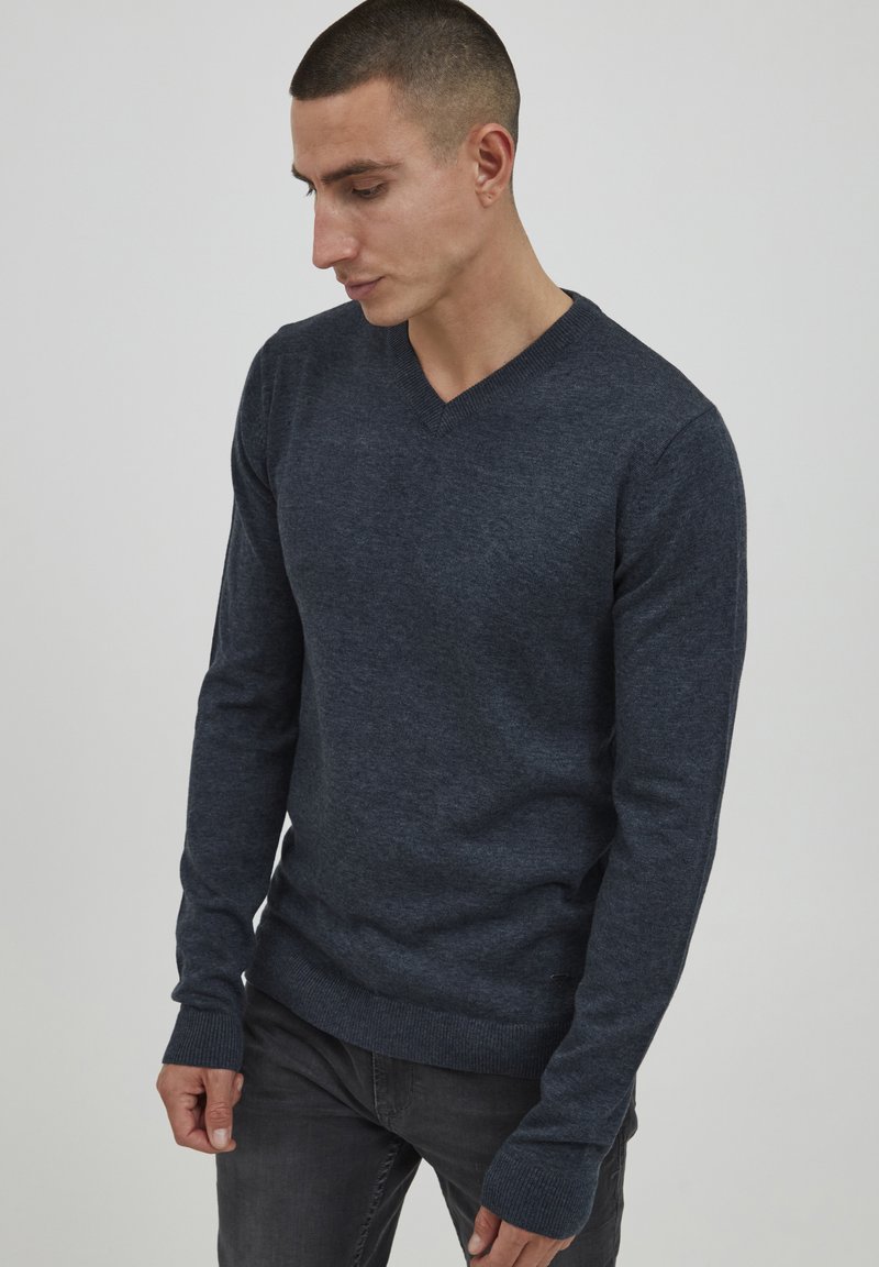 Maglione blu a scollo a v realizzato in tessuto lavorato a maglia con maniche lunghe e rifiniture a coste. Indossato con pantaloni grigio scuro, tenuto da una persona di lato.