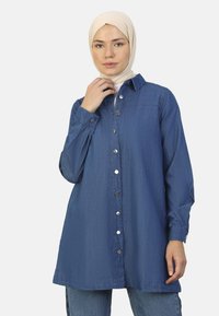 Camisa de mezclilla de botones en azul profundo, con cuello, mangas largas y botones de presión plateados en la parte delantera. Textura suave.