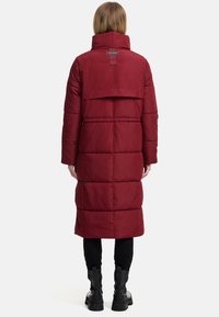 Lange bordeauxrode pufferjas met een hoge kraag, horizontale stikdetails en een achterste vent. Draag een zwarte broek en stevige laarzen.