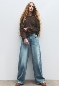 Rjava dolga majica z dolgimi rokavi, svetlo pranih širokih hlačah in rjavih loaferjih. Lase ima dolge in valovite; stoji ob belem ozadju.