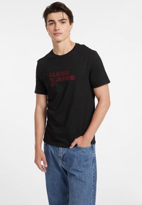 T-shirt en coton noir à manches courtes et col rond, arborant un logo rouge "GUESS JEANS" sur le devant. Porté avec un jean en denim bleu.