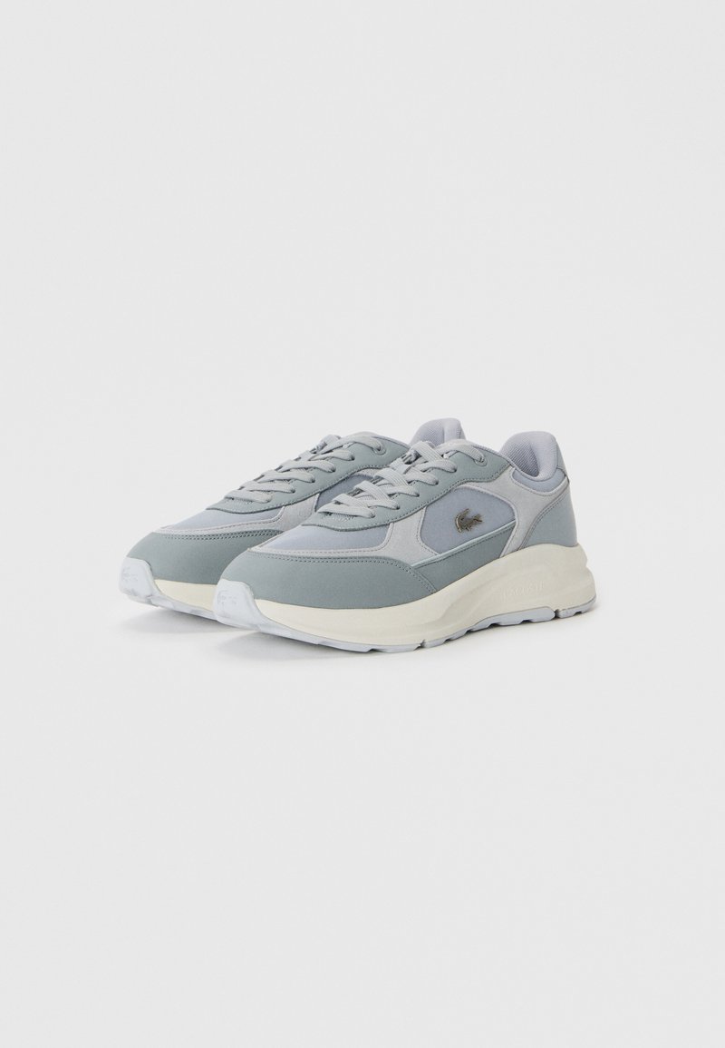 Une paire de baskets grises et blanches à lacets avec des semelles épaisses et un petit logo brodé sur le côté, placée sur un fond blanc.