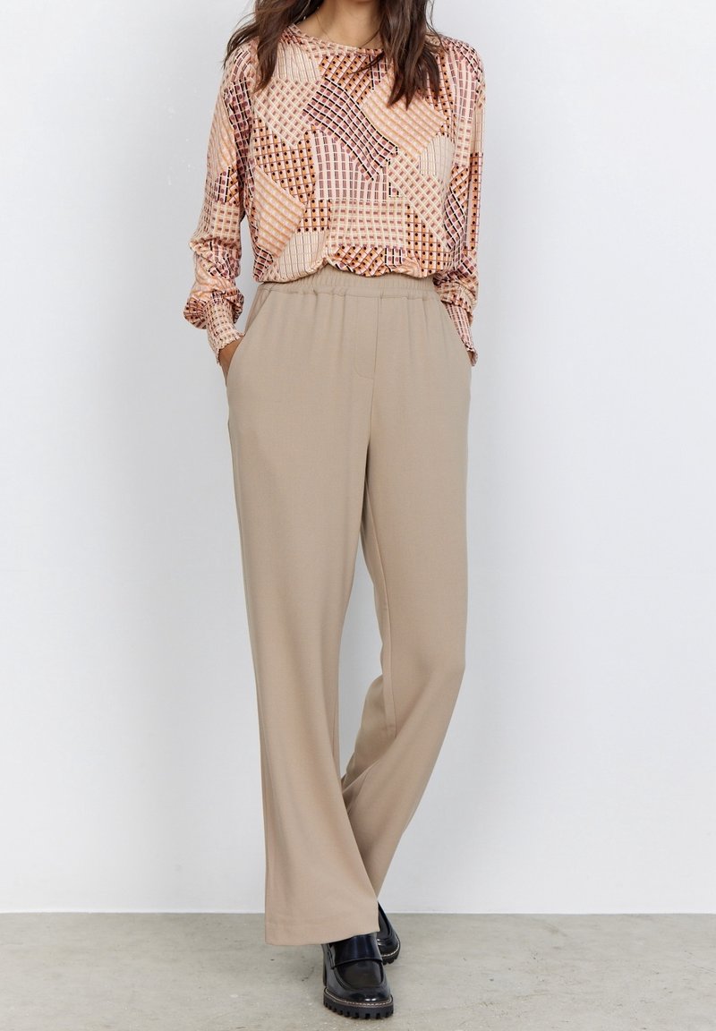 Long-sleeve top with a geometric pattern in warm tones; beige wide-leg trousers. Model wears black platform shoes. Neutral backdrop.