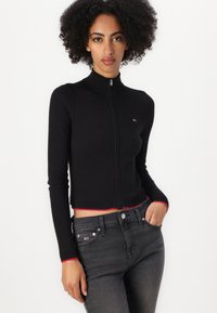 Suéter negro de canalé con cremallera y un alto cuello, con un acento rojo en el dobladillo. Presenta mangas largas y un diseño ajustado, combinado con jeans grises.