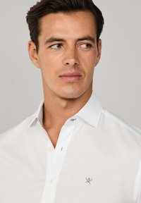 Man in een wit overhemd met een klein logo op de borst, kijkend naar rechts voor een effen achtergrond.