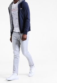 Námořnická mikina s kapucí a pruhovanou podšívkou, bílý tričko s grafickým potiskem se horizontálními pruhy, světle šedé džíny a bílé tenisky.