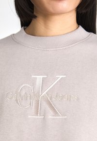 Svijetlosivi dukserica s rebrastim izrezom i vezom "CK" logotipa s natpisom "Calvin Klein Jeans" ispod u suptilnom sjaju.