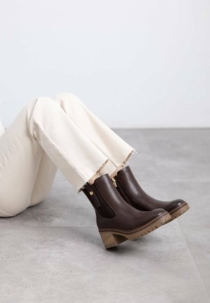 Botines de cuero marrón con cremalleras laterales, tacones apilados y suelas de goma texturizadas, combinados con pantalones de color claro y dobladillo deshilachado.