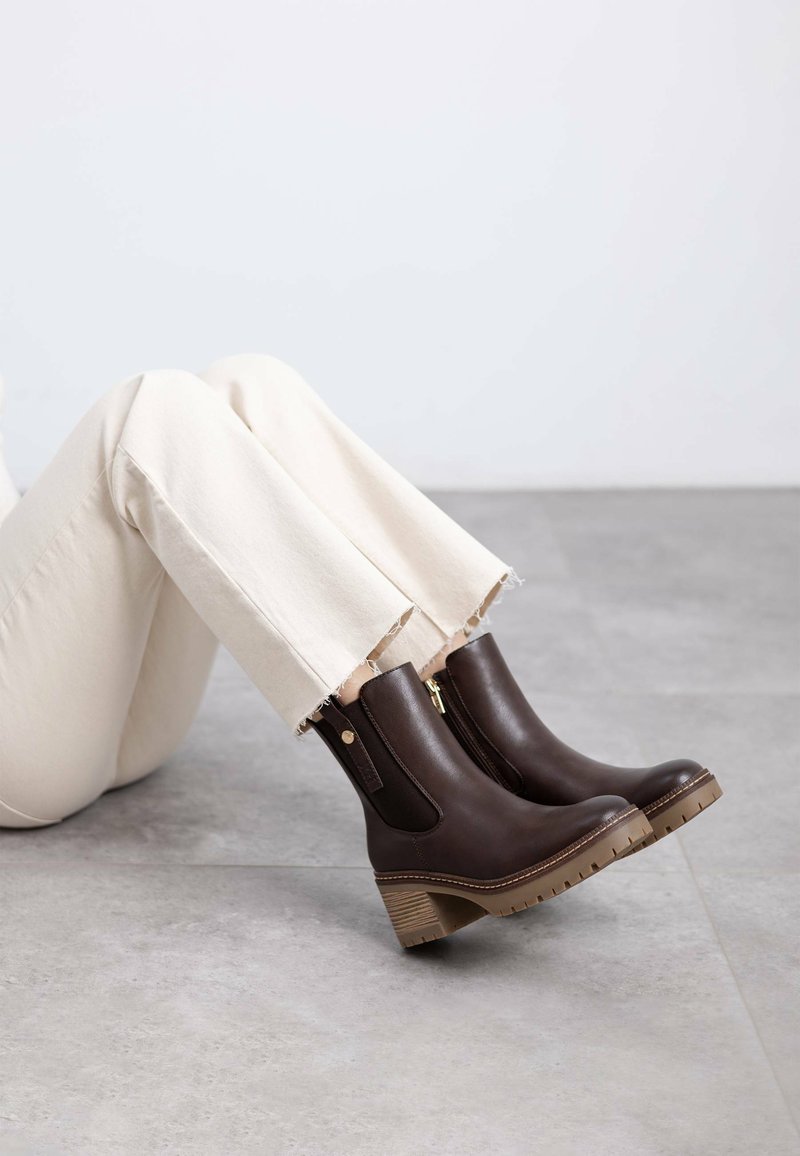 Botines de cuero marrón con cremalleras laterales, tacones apilados y suelas de goma texturizadas, combinados con pantalones de color claro y dobladillo deshilachado.