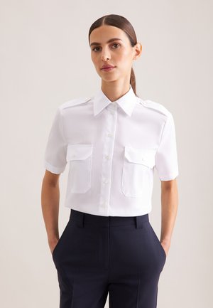 Donna con capelli scuri raccolti indietro indossa una camicia bianca e stirata a maniche corte con bottoni e due tasche frontali, e pantaloni scuri a vita alta.