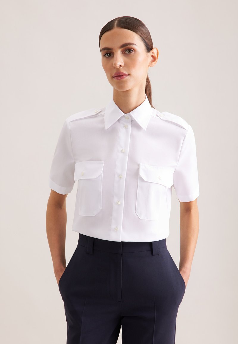 Femme aux cheveux foncés attachés en arrière porte une chemise blanche nette à manches courtes avec deux poches avant et un pantalon sombre taille haute.