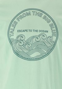 Grön bomull t-shirt med en cirkulär grafik som har vågor och texten: "Berättelser från Stora Blå" och "Rym till Havet."