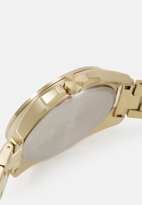 Reloj de tono dorado con una caja redonda, bisel texturizado y una corona prominente. La correa presenta un acabado pulido, mostrando eslabones y un diseño elegante.