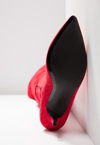 Botte à talon haut en suède rouge avec un bout pointu et une semelle en caoutchouc noire texturée. Le dos est orné d'une fermeture éclair.