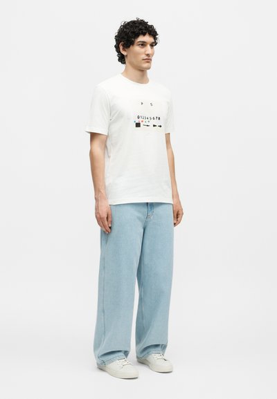 Άνδρας με κυματιστά σκούρα μαλλιά, που φοράει λευκό μπλουζάκι με γραφικά, ανοιχτό μπλε τζιν παντελόνι με φαρδιά μπατζάκια και λευκά αθλητικά παπούτσια, στέκεται μπροστά σε απλό φόντο.