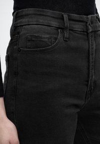 Κοντινό πλάνο ενός ατόμου που φοράει μαύρο τζιν με μπροστινή τσέπη και θηλιές για ζώνη, συνδυασμένο με μαύρο τοπ.