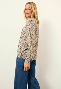 Blonde woman in floral long-sleeve blouse and blue jeans standing sideways, looking over shoulder against a plain light background.