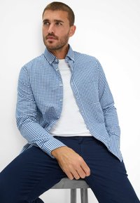 Camicia a quadri blu e bianchi in tessuto leggero, con colletto button-down, maniche lunghe, indossata sopra una t-shirt bianca, abbinata a pantaloni blu navy.