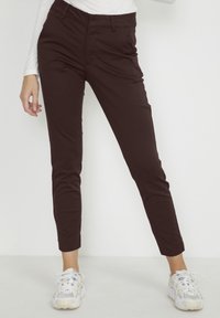 Tmavě hnědé skinny kalhoty s hladkou texturou, s mírným vzestupem v pase a slim fit až k kotníkům. Vydané s bílými teniskami.