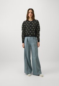 Black knitted cardigan with gold geometric pattern, button front, and balloon sleeves, paired with light blue wide-leg trousers.