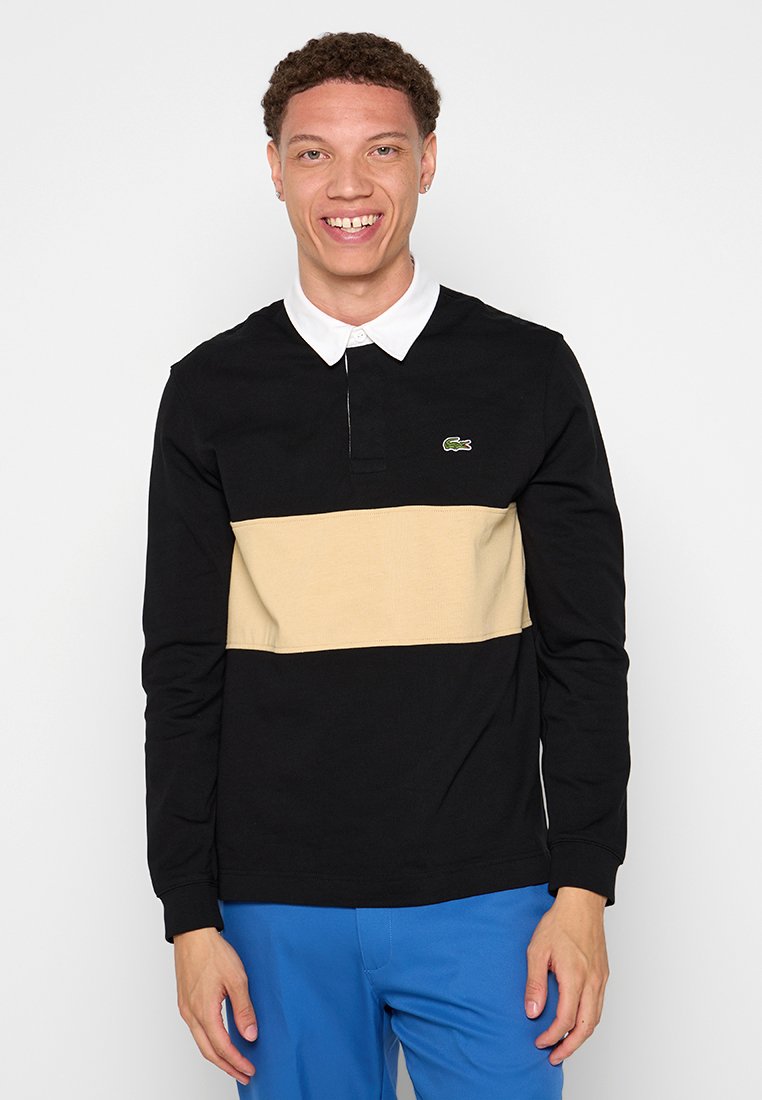 Polo noir à manches longues avec une rayure horizontale beige, un col blanc et un accent logo vert. Matériau en coton, coupe régulière, design décontracté.