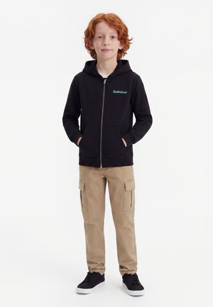Dieťa s červenými vlasmi stojí s rukami v vreckách, oblečené v čiernej mikine Quiksilver, khaki cargo nohaviciach a čiernych teniskách na bielom pozadí.