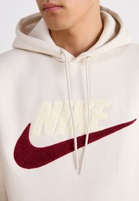 Gräddfärgad hoodie med en framficka, prydd med en strukturerad beige "NIKE"-logotyp och ett vinrött Swoosh-emblem. Inkluderar justerbara dragsnören.