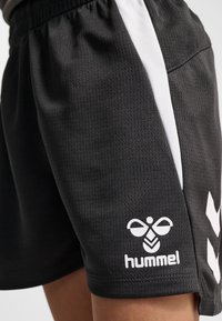 Sorte atletikshorts med hvid sidepanel og Hummel-logo på den nederste lårdel, båret af en person mod en ensfarvet baggrund.