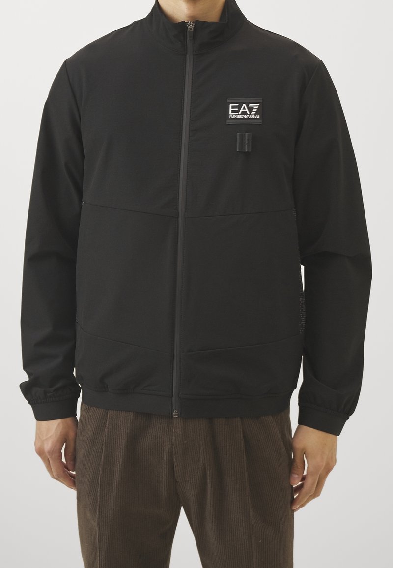 Svart zip-up jacka i lätt tyg, med en bröstlogotyp, elastiska ärmslut och sidofickor med en texturerad yta.