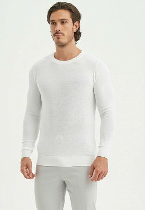 Chemise tricotée blanche à manches longues avec un col rond et des poignets côtelés, présentant un tissu texturé et une coupe ajustée.