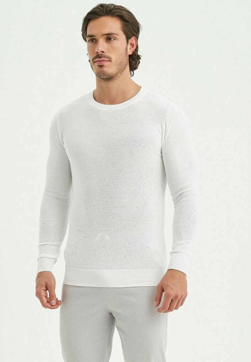 Chemise tricotée blanche à manches longues avec un col rond et des poignets côtelés, présentant un tissu texturé et une coupe ajustée.