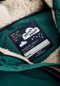 Štítok nepremokavej bundy vo vnútri zelenej látky s krémovým flísovým podšívaním, zobrazujúci pokyny na starostlivosť a miesto na meno.