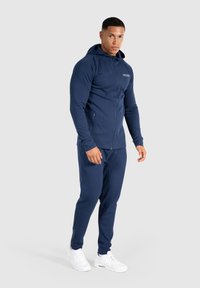 Felpa con zip color navy dal design aderente, abbinata a pantaloni coordinati. Realizzata in tessuto morbido, presenta una tasca a marsupio e scarpe da ginnastica bianche.