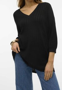 Nainen, jolla on päällään löysä musta V-neck-neule, jossa on kolme neljännestä hihaa, siniset farkut ja suuri kultaketju vasemmassa ranteessaan.