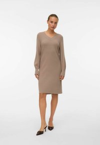 Robe tricotée en beige clair avec un décolleté en V, des manches longues et une silhouette droite, accompagnée de talons aiguilles marron. Texture lisse.