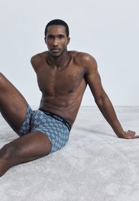 Cuecas tipo boxer masculinas com padrão geométrico azul e cós preto com a inscrição "HUGO", feitas em tecido macio e elástico para maior conforto.