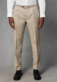 Pantalon beige plissé à la texture lisse, doté d'une taille avec bouton et passants de ceinture, assorti à des chaussures noires.