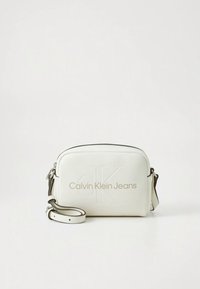 Witte leren crossbodytas met een afgeronde vorm, voorzien van een geperst "CK"-logo en de tekst "Calvin Klein Jeans" in goud. Verstelbare band inbegrepen.