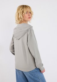 Lys grå hettegenser med avslappet passform, med snøring i hetten, lange ermer og en jevn tekstur. Kombinert med løse blå jeans.
