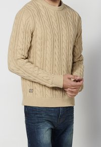 Pull en maille beige avec un motif torsadé, col rond, poignets et ourlet côtelés, porté avec un jean foncé. Petite étiquette sur le côt é.